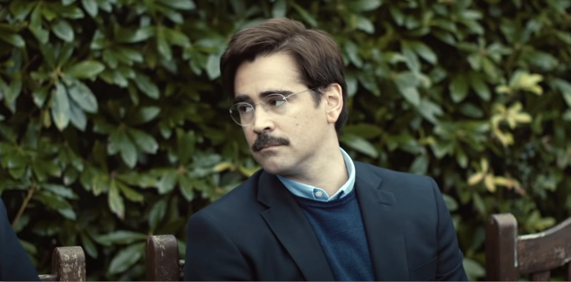 Colin Farrell in a scene from "The Lobster," wearing glasses, a dark blazer, and a blue sweater, seated on a bench with greenery in the background
