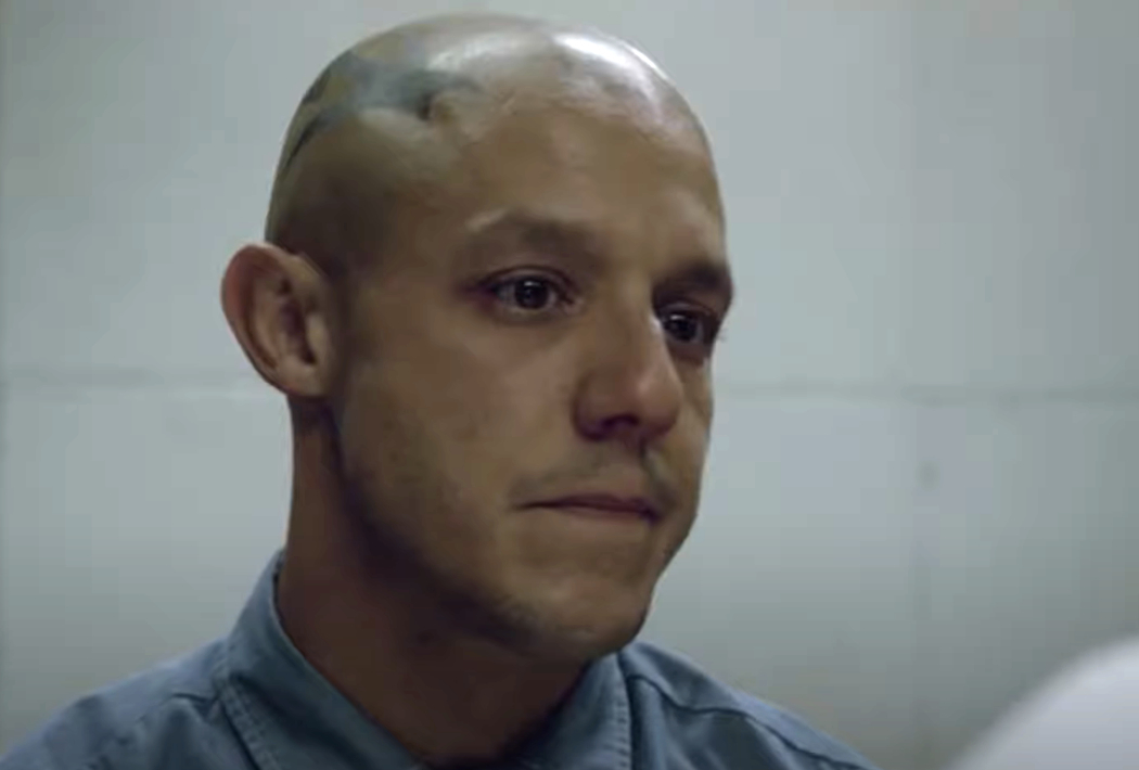 A solemn man with a shaved head and a tattoo on his scalp looks off-camera in a somber scene, wearing a button-up shirt