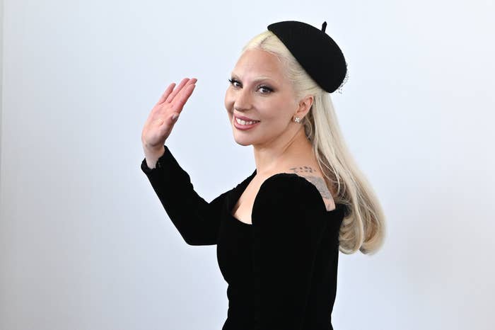 Lady Gaga smiling and waving, dressed in a dark long-sleeve outfit with a matching beret