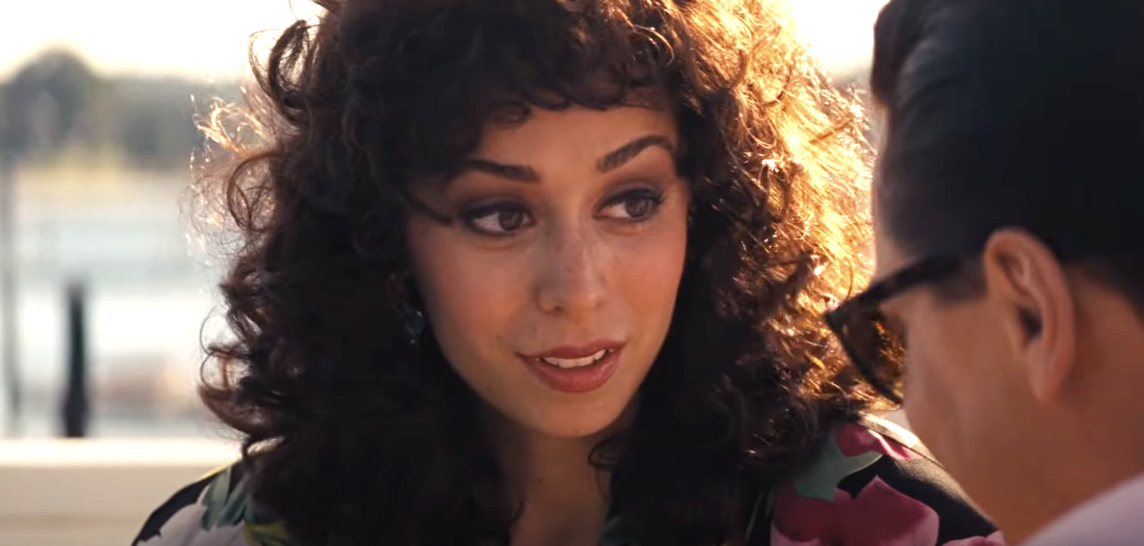 A woman with curly hair and a floral-patterned outfit engages in a conversation with a man wearing glasses, in a scene from a movie or TV show