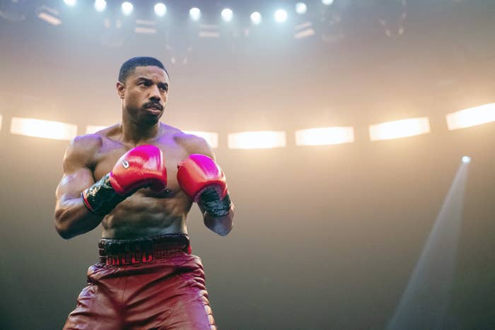 Michael B. Jordan in a boxing stance with red gloves and boxing shorts, under bright circular lights