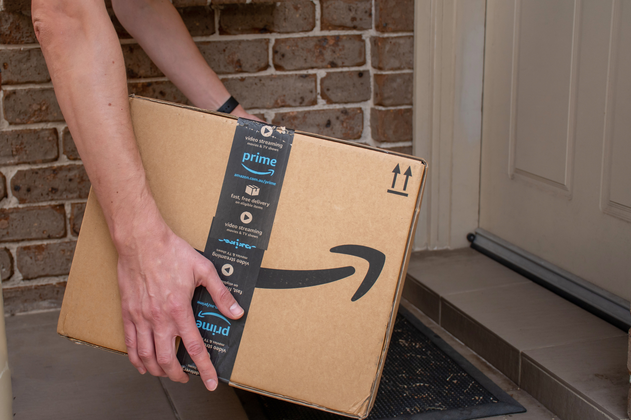 A person's hands are seen placing an Amazon Prime package on the doorstep of a house