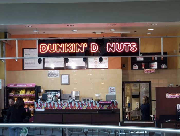 Inside view of a Dunkin' Donuts store with customers ordering. The illuminated sign reads "Dunkin' D NUTS" due to a missing letter