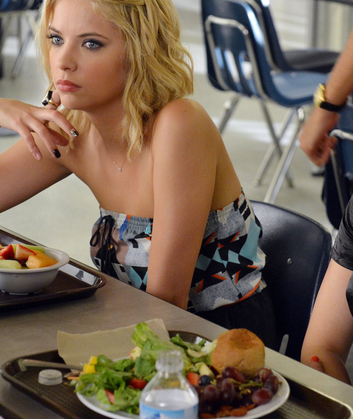 Ashley Benson sits at a cafeteria table with lunch trays and food, while chatting with others out of frame. She has shoulder-length blonde hair