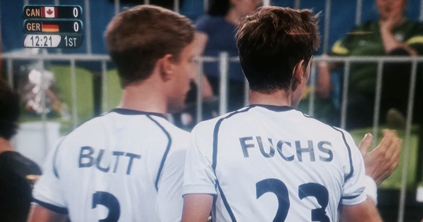 Butt and Fuchs, Canadian and German soccer players, respectively, during a match shown on the scoreboard, stand on the field with "12:21" in the first half