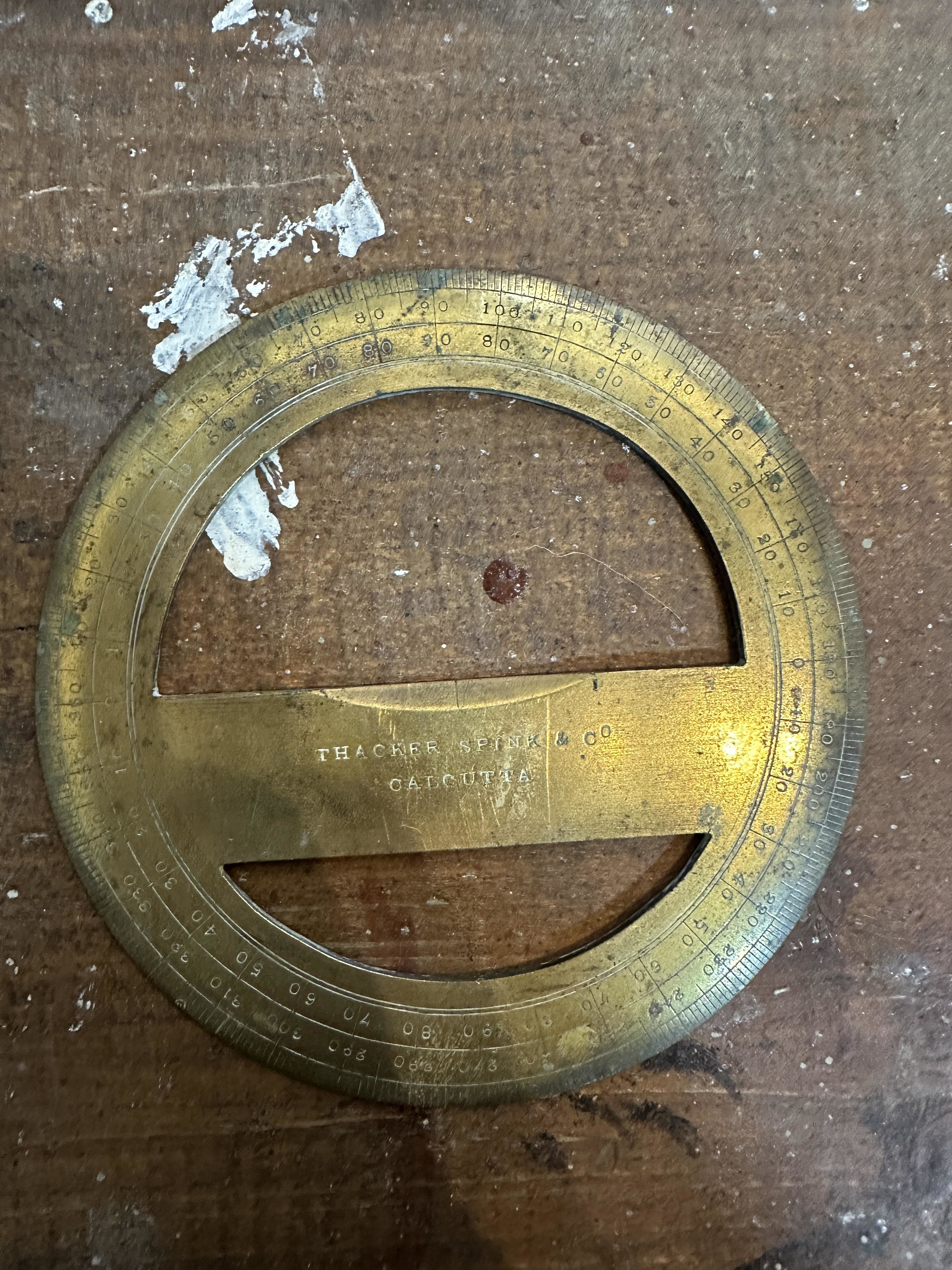 A circular brass protractor with a central cut-out, inscribed "Thacker, Spink &amp;amp; Co, Calcutta," placed on a wooden surface