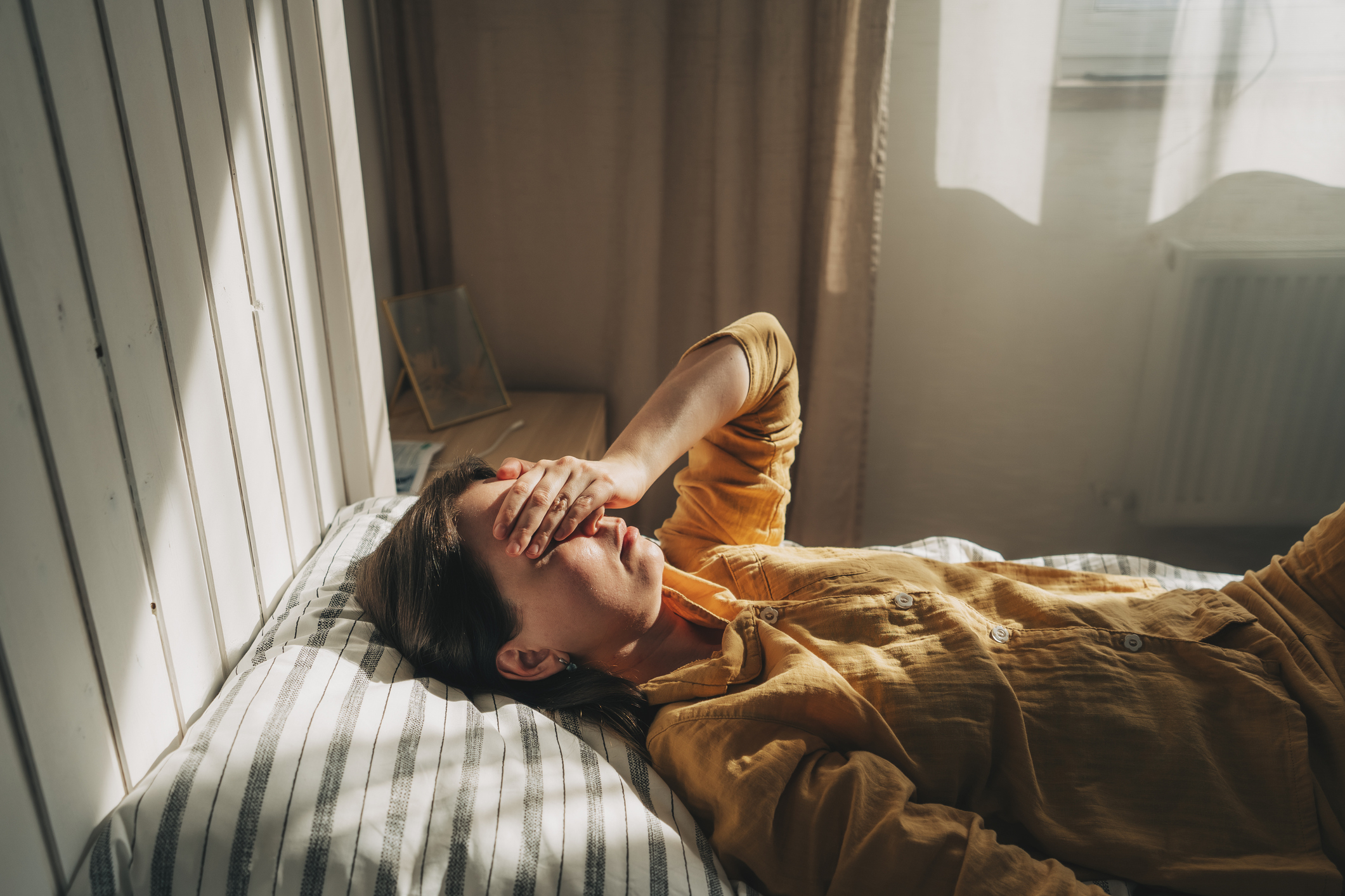 A person lying in bed wearing a comfortable outfit, with one hand covering their face, appearing stressed or tired