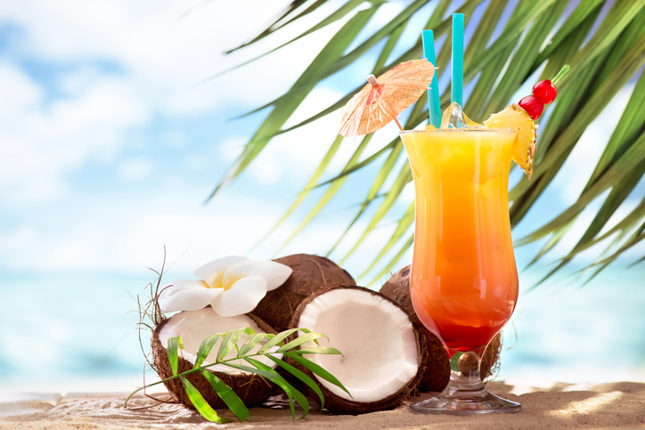 Tropical beach scene with a cocktail garnished with a mini umbrella, pineapple, and cherries in a glass, surrounded by coconuts, a white flower, and palm leaves