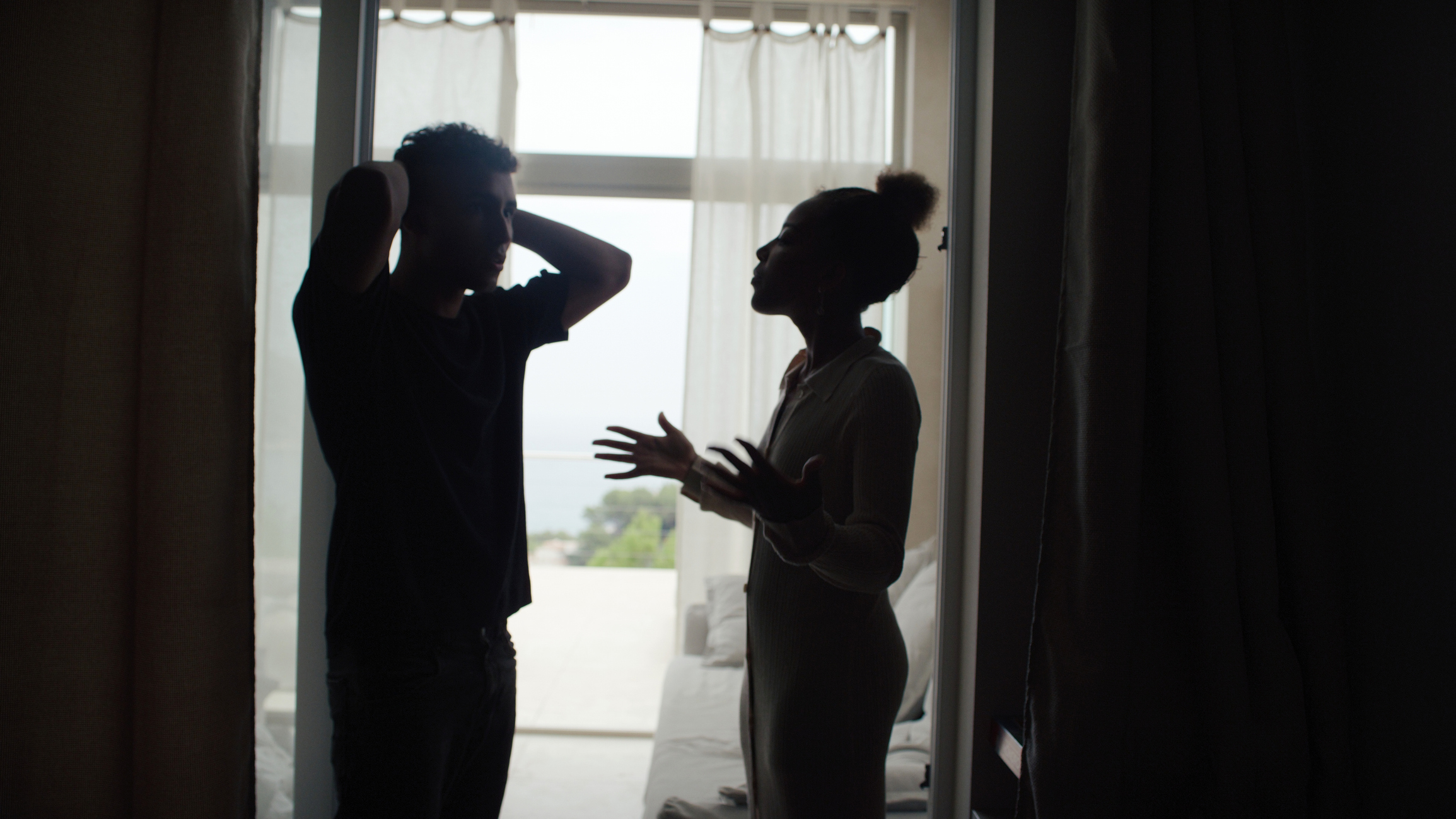 A man and woman stand indoors, silhouetted against a window; the man has his arms raised behind his head, and the woman gestures animatedly while speaking