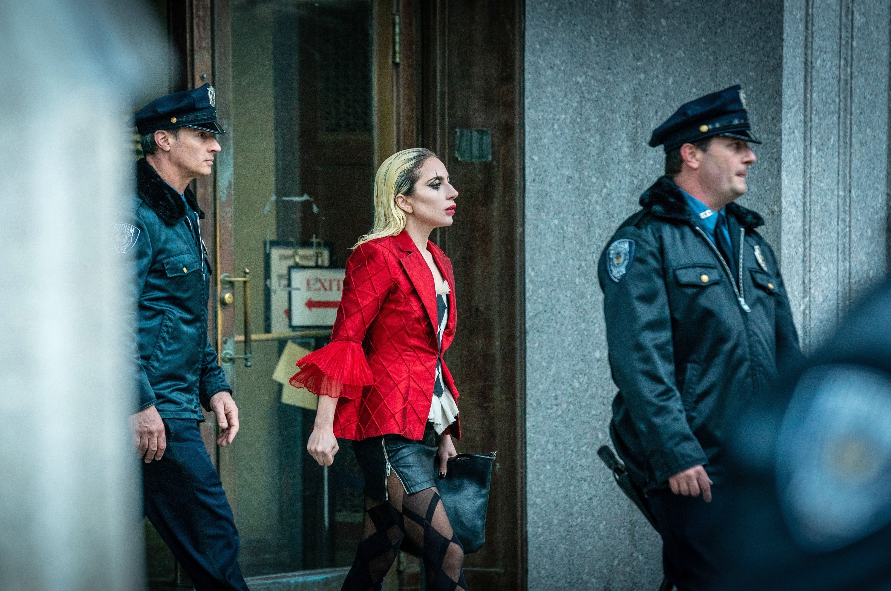 Lady Gaga, in a jacket with dramatic sleeves and black leather pants, walks out of a building escorted by two police officers in uniform in a scene from the film