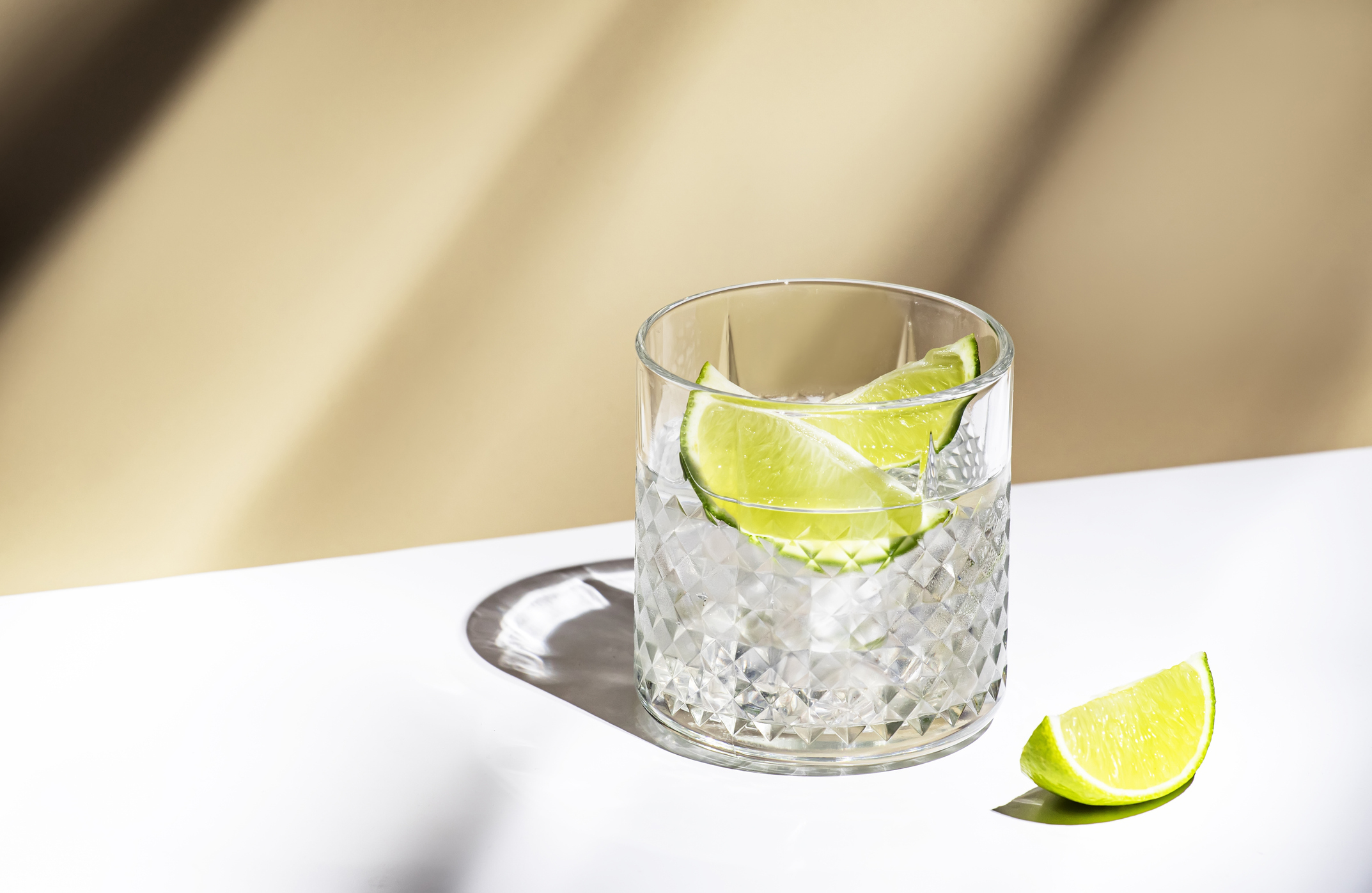 A glass filled with ice and lime slices sits on a white surface with a lime wedge beside it. Shadows fall across the background