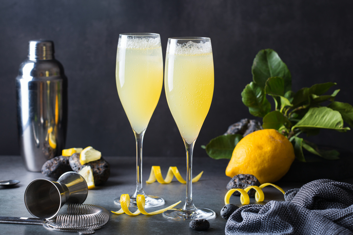 Two filled champagne flutes with lemon peels, a metal shaker, a lemon, and a strainer on a table