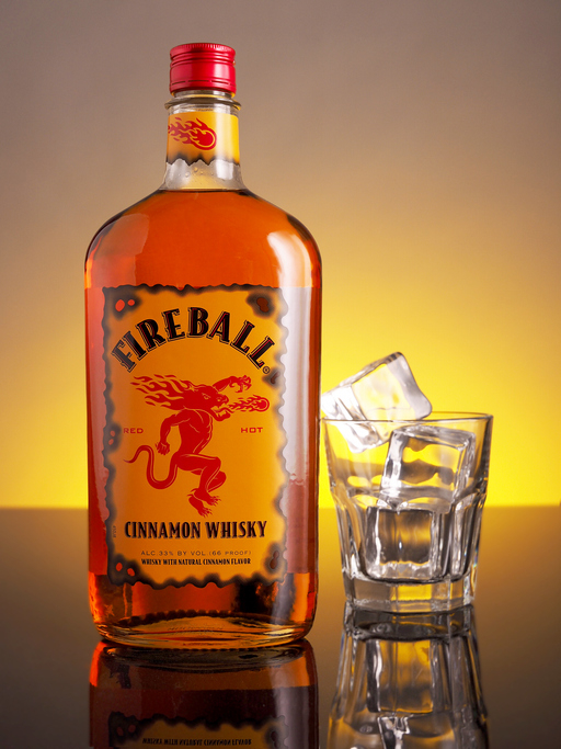 Fireball Cinnamon Whisky bottle next to a glass with ice cubes, against a glowing background