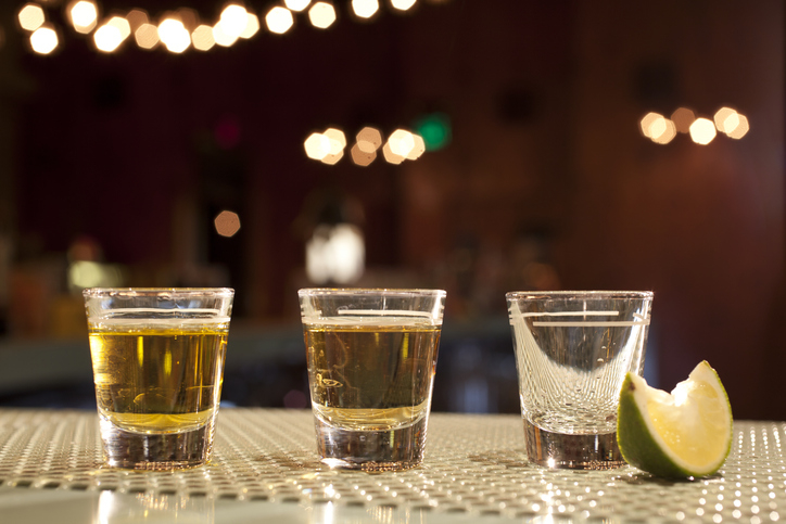 Three shot glasses are lined up on a bar counter; two contain liquid, while one is empty. A lime wedge is placed next to the empty glass