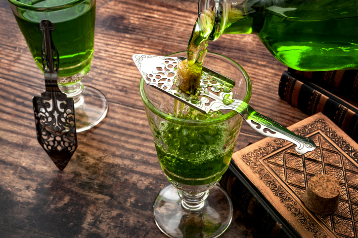 Absinthe being poured into a glass through a perforated spoon holding a sugar cube, with another glass and books in the background