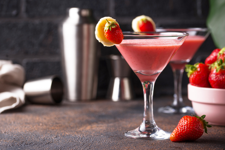 Two strawberry daiquiris garnished with strawberries and banana slices, placed on a counter with a cocktail shaker and fresh strawberries in the background