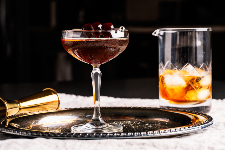 A cocktail glass filled with a dark liquid, garnished with cherries, on a silver tray with a gold jigger and a beaker containing an orange drink with ice in the background