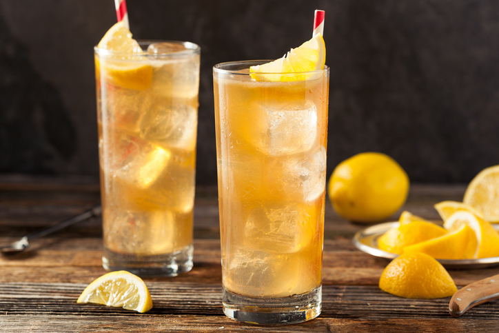 Two tall glasses of iced tea with lemon wedges and striped straws, placed on a wooden table, with lemon slices in the background
