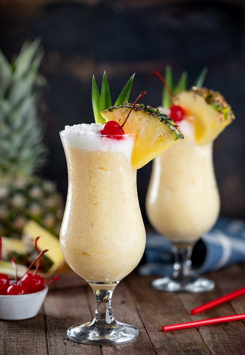 Two Pina Colada cocktails garnished with a cherry, pineapple slice, and leaf. One is in the foreground; the other is blurred in the background