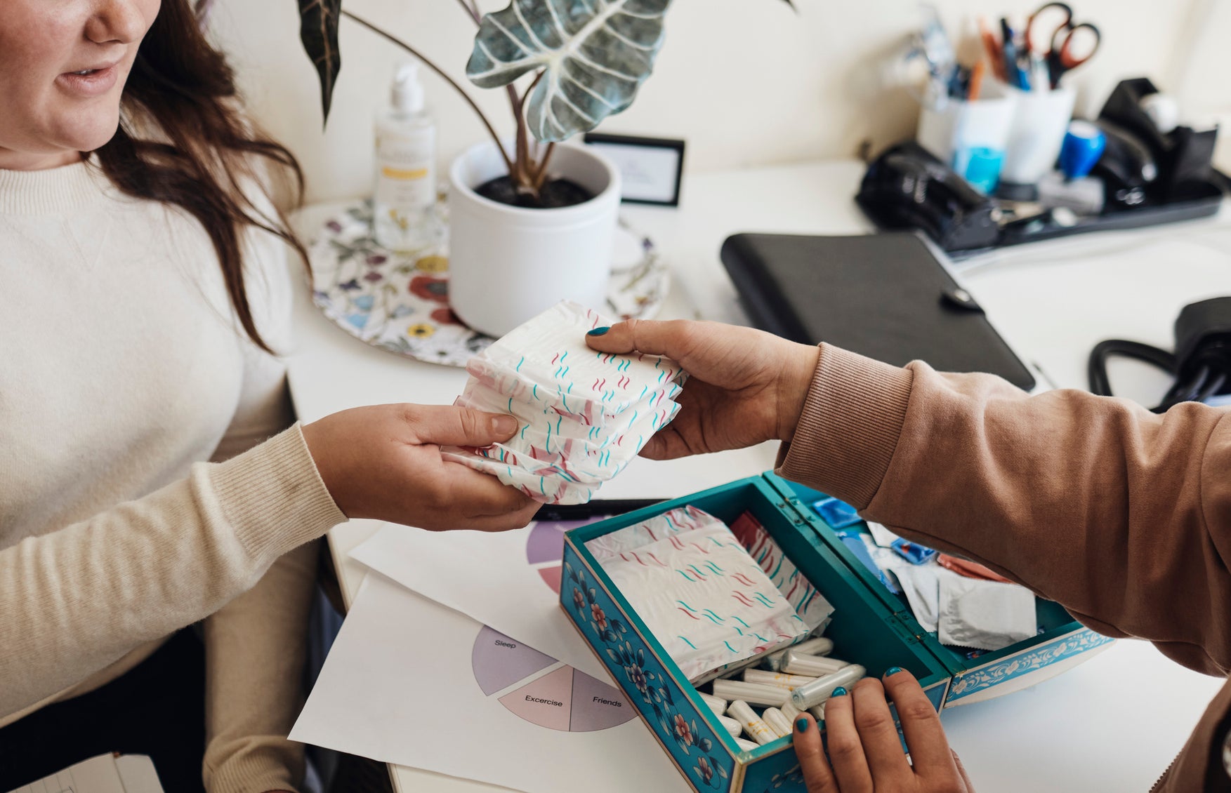 Two women sit at a desk discussing and handling feminine hygiene products from a box. A potted plant and office supplies are in the background
