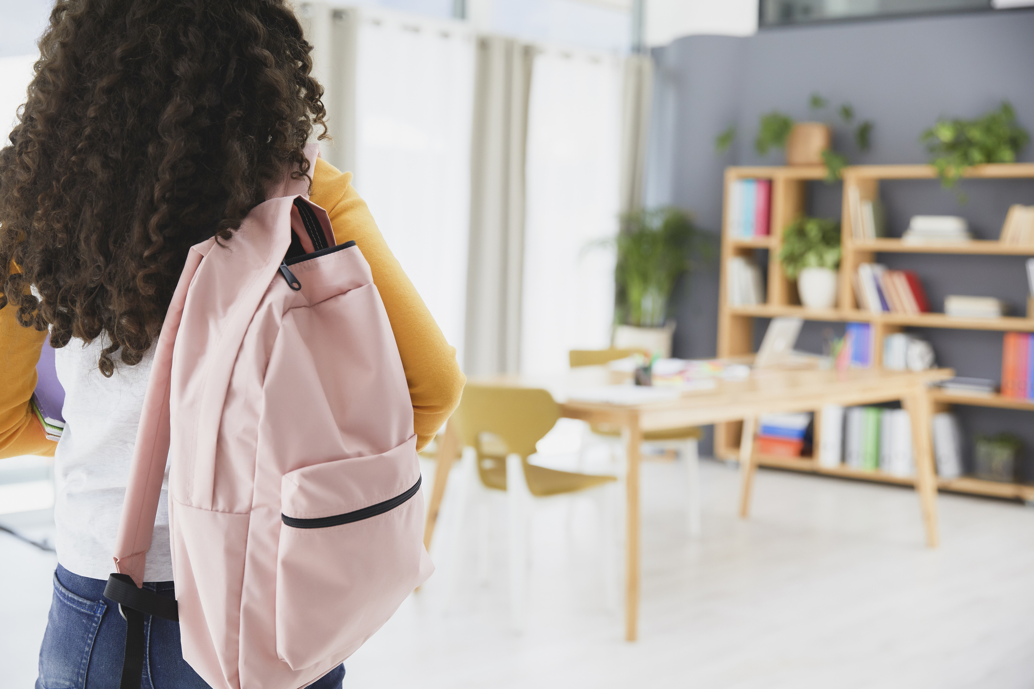 A person with curly hair and a backpack is standing in a bright, well-organized library with bookshelves and a large table