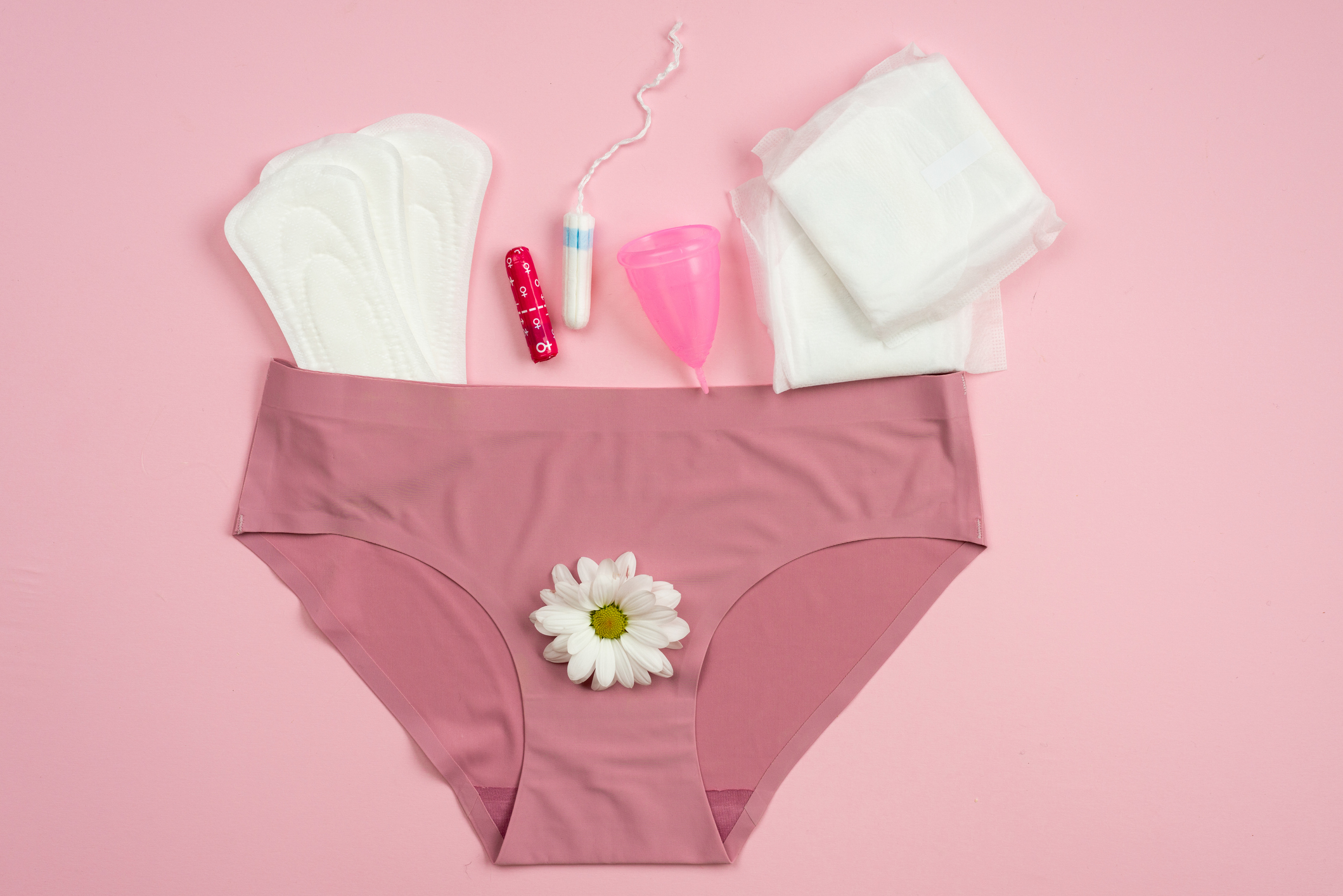 Various menstrual products are arranged around a pink pair of underwear with a daisy on them, showcasing options like pads, tampons, and a menstrual cup