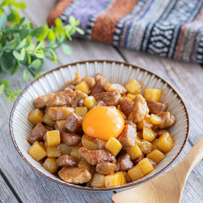 肉と野菜が入った丼の上に生卵が載せられている料理。背景には布と観葉植物が置かれています。