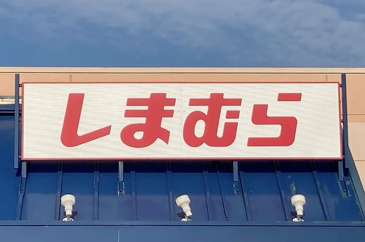 A store sign with Japanese characters reads &ldquo;しまむら&rdquo; above the entrance. The building has a partially visible blue awning and a clear sky above