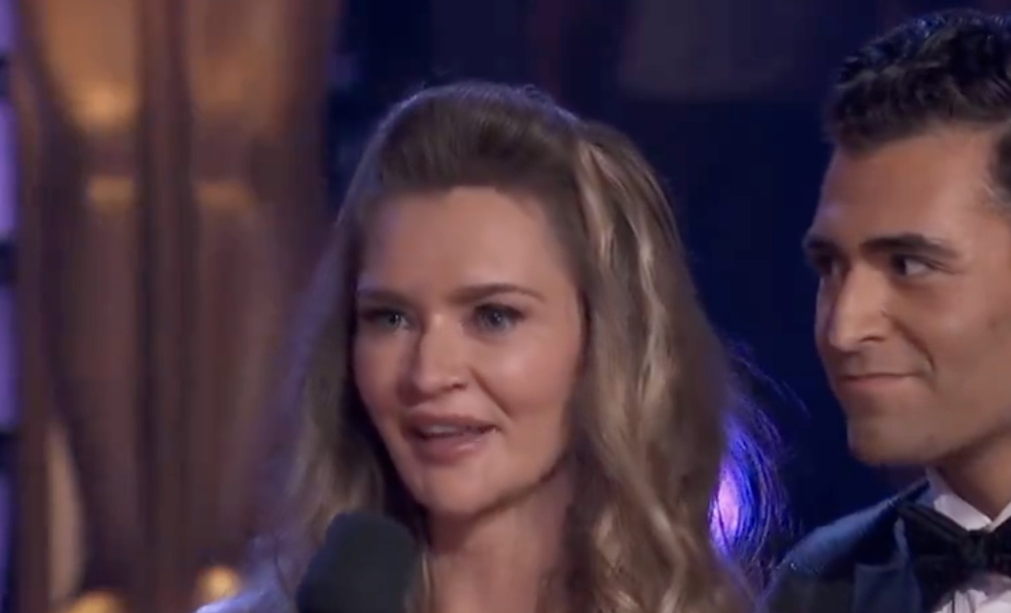 A woman with long, wavy hair and a man in a tuxedo are being interviewed. They are in a formal setting, possibly at an event or show