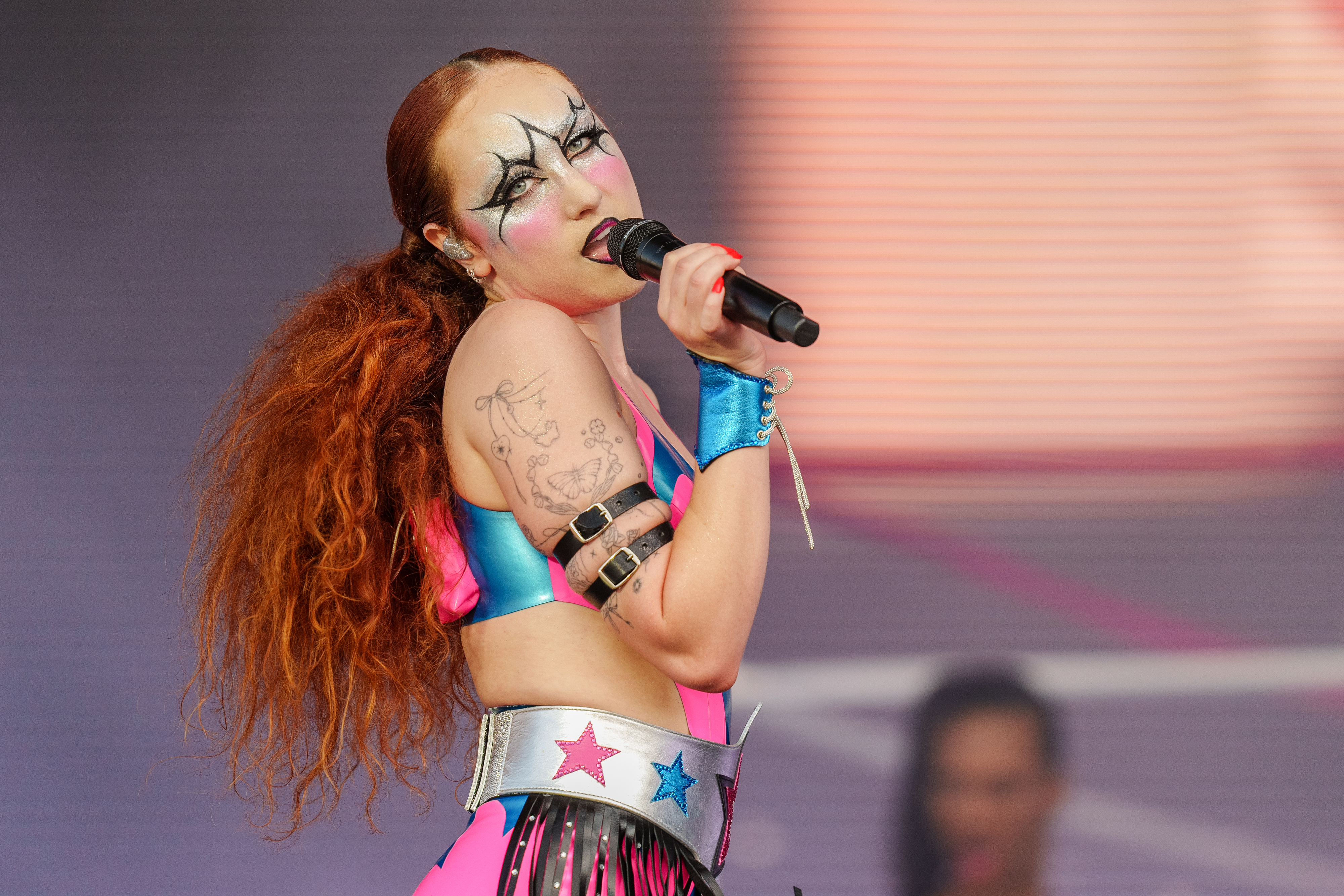 Chappell Roan performing on stage, wearing a colorful futuristic outfit with star designs, bold face makeup, and holding a microphone