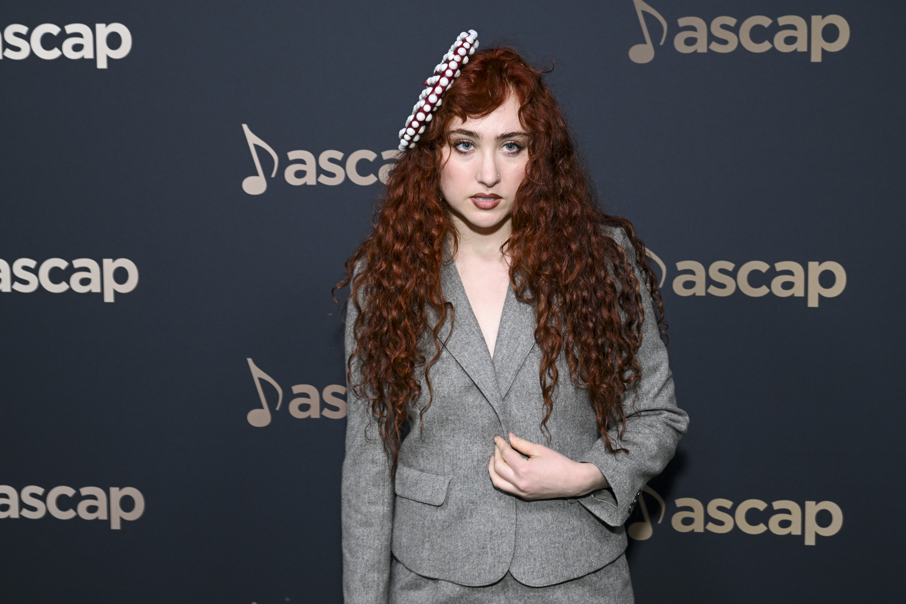 Chappell Roan with long curly red hair, dressed in a suit, stands in front of an ASCAP backdrop. She is wearing a hat decorated with pearls