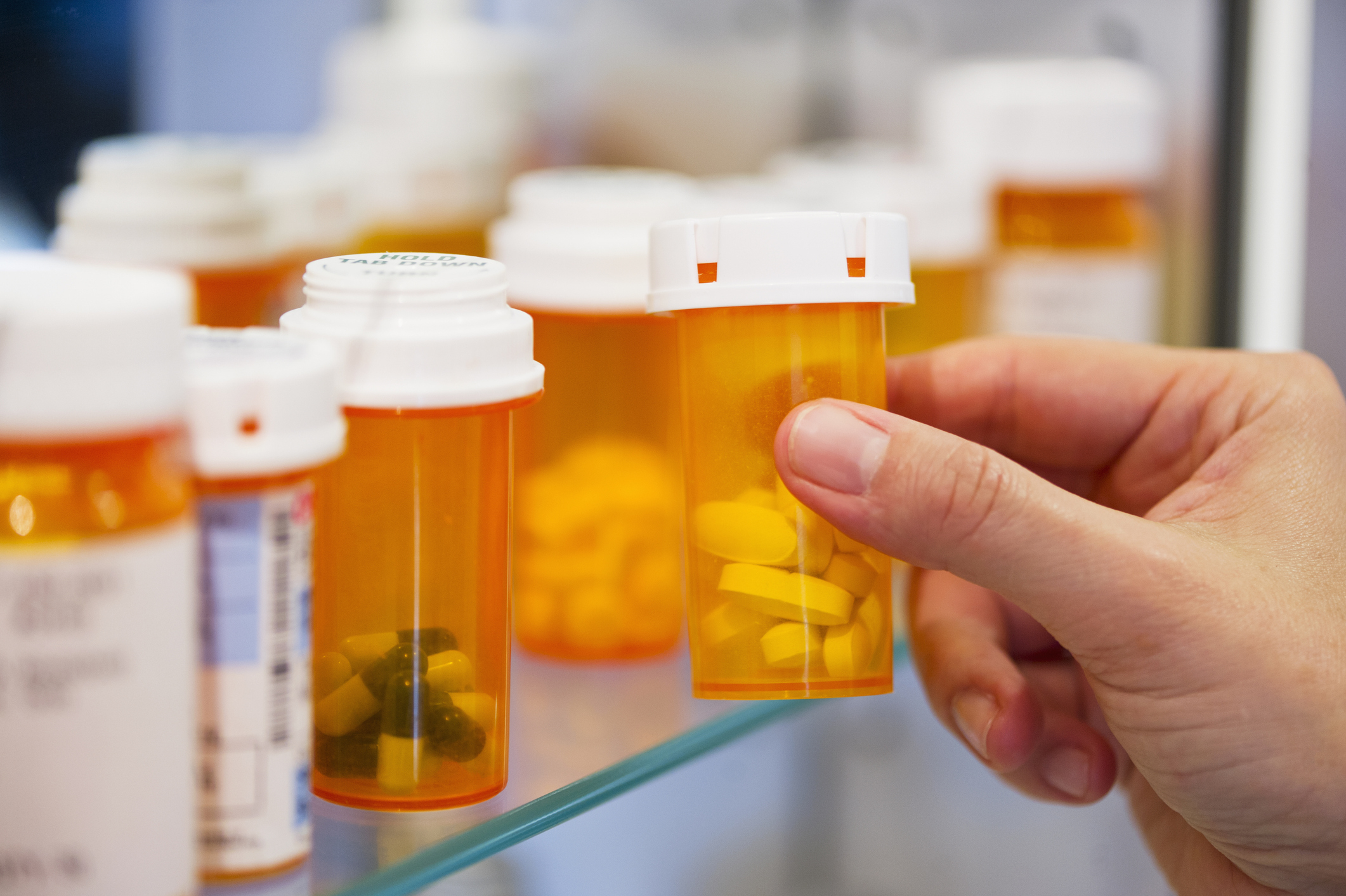 A hand reaches for a prescription pill bottle on a shelf filled with various other prescription pill bottles