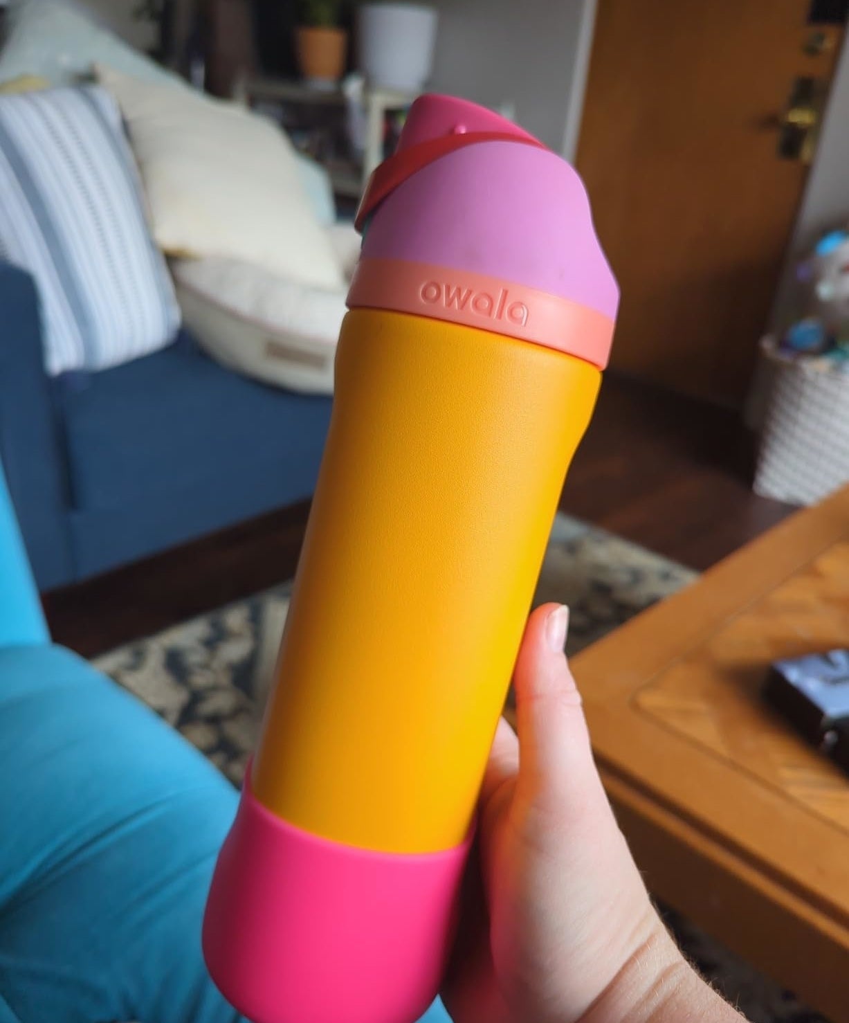 A hand is holding a vibrant Owala water bottle with a pink and orange design in a cozy living room