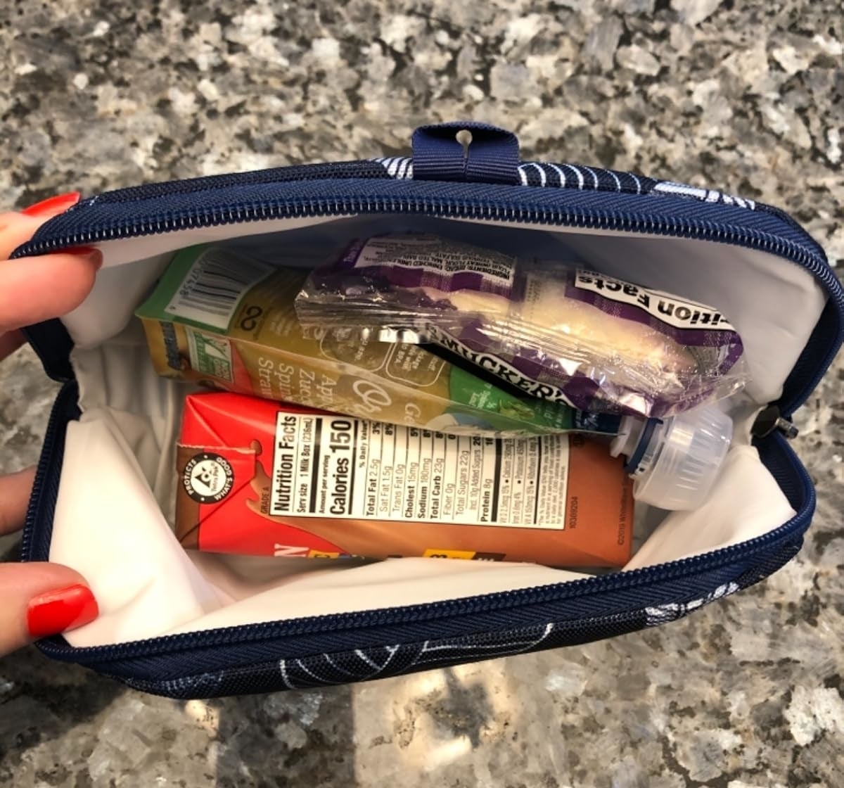 A hand holds an open pouch containing snacks: a juice box, granola bar, raisins, and a tiny water bottle