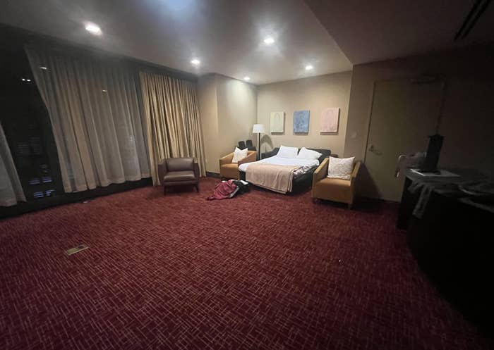A hotel room with dim lighting, featuring a bed, nightstands, a sofa, an armchair, a desk, and several bags and clothes on the floor