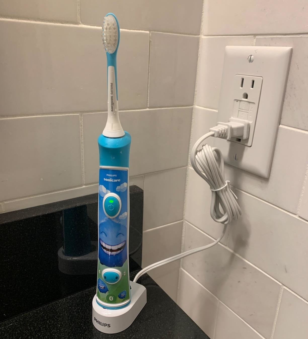 Kids' Philips Sonicare electric toothbrush on a charging stand, plugged into an outlet in a bathroom. The toothbrush features a smiling face design