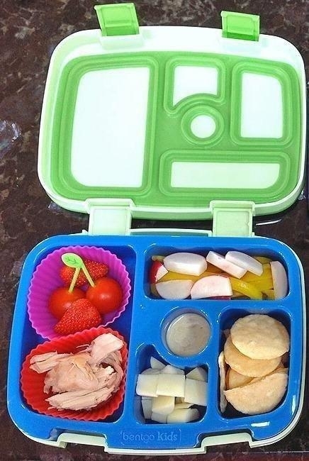 Open bento lunchbox with compartments containing cherry tomatoes, strawberries, cheese cubes, crackers, turkey slices, radish slices, and dip