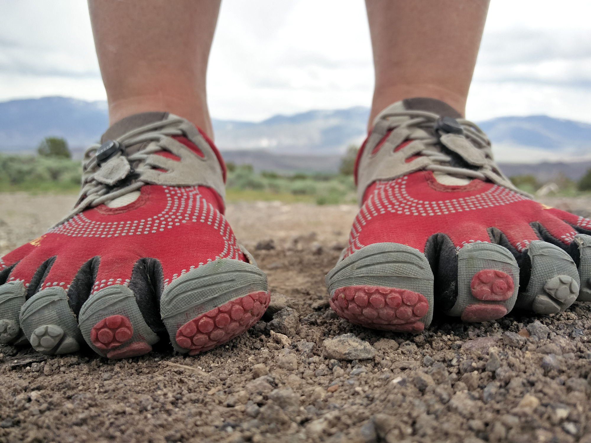 Close-up of a person wearing unique five-finger running shoes on a rocky trail with mountains in the background