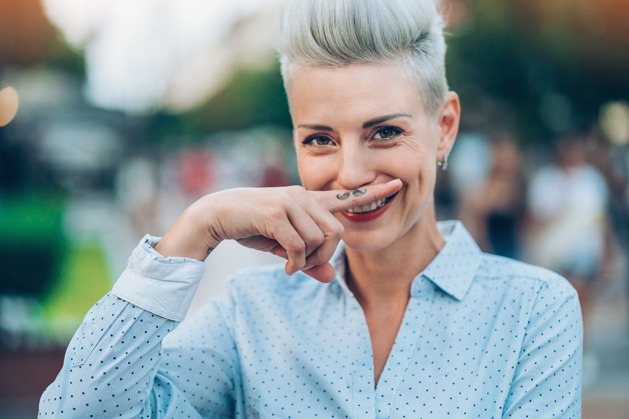 Person with a short, stylish haircut and polka dot shirt playfully holds their index finger with a tattoo of a mustache under their nose, smiling
