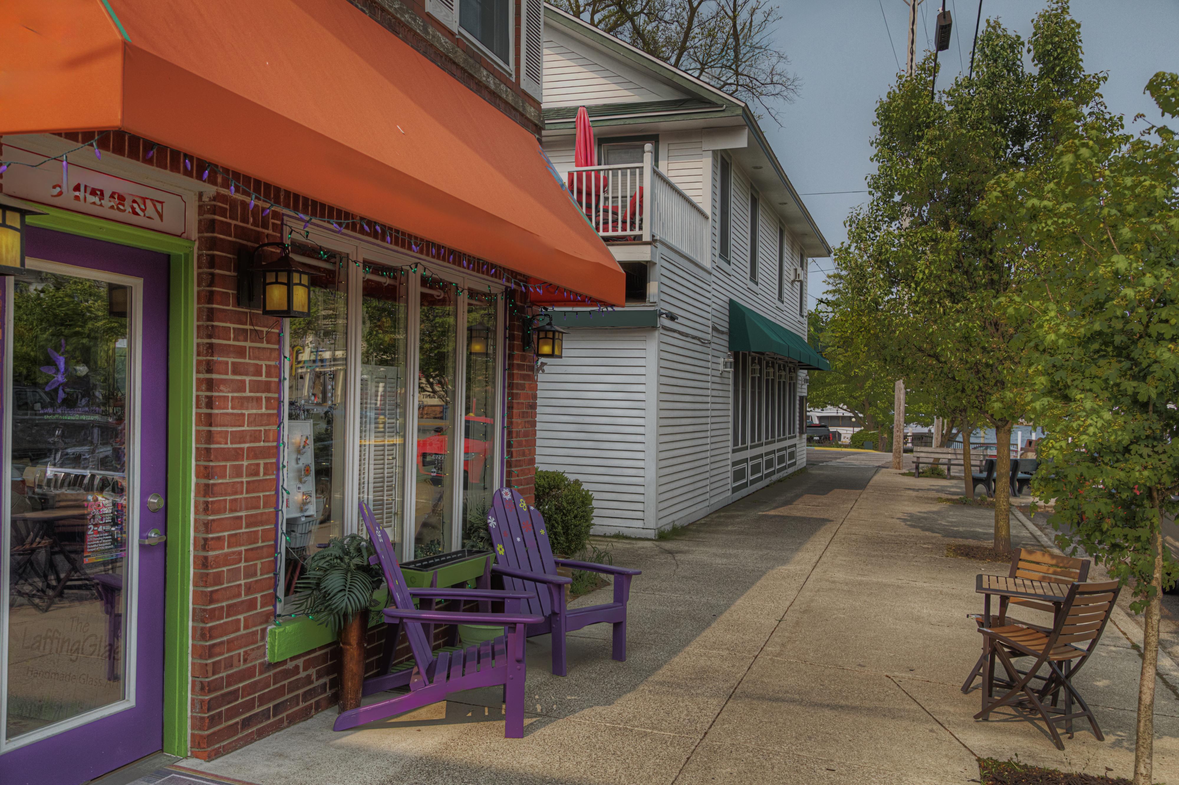 Sidewalk in a small town with a bright storefront featuring orange awnings and purple chairs on the left, and a white building with green awnings further down the street