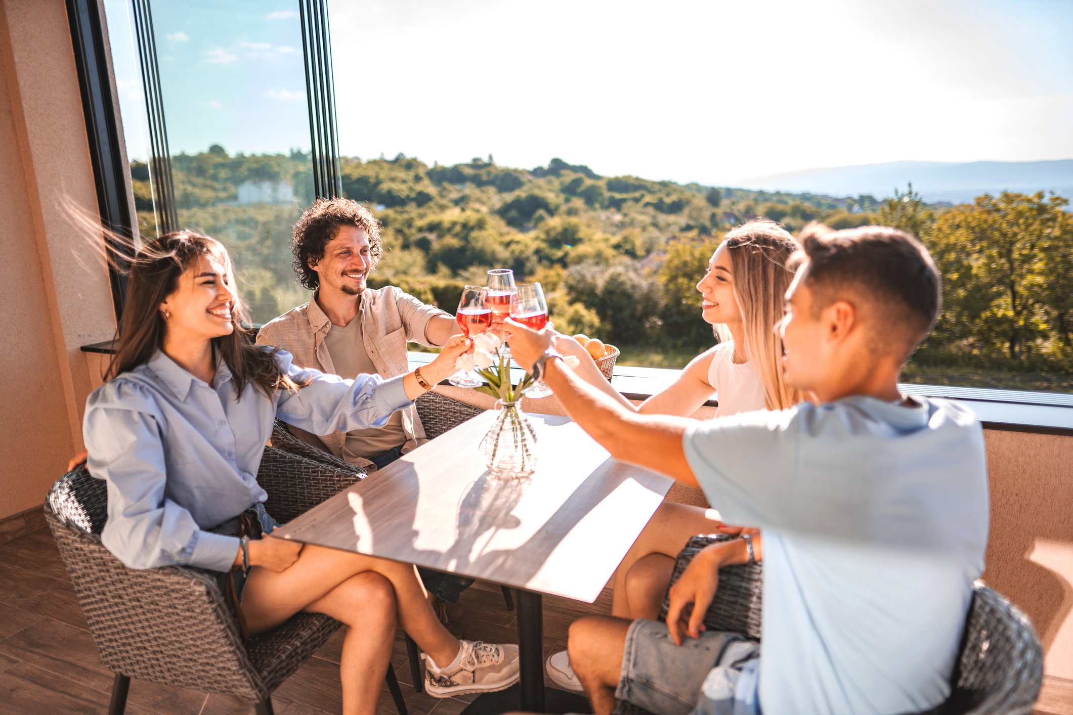 Four people enjoying a meal together at an outdoor restaurant, cheerfully clinking their glasses. The background features a scenic view of greenery and hills