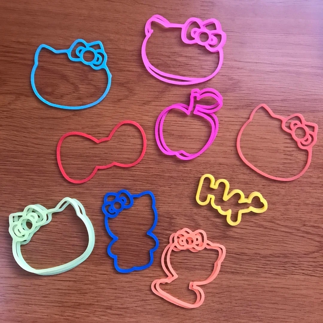 Nine colorful rubber bands shaped like Hello Kitty faces, a bow, an apple, "HK", and other playful designs, arranged on a wooden surface