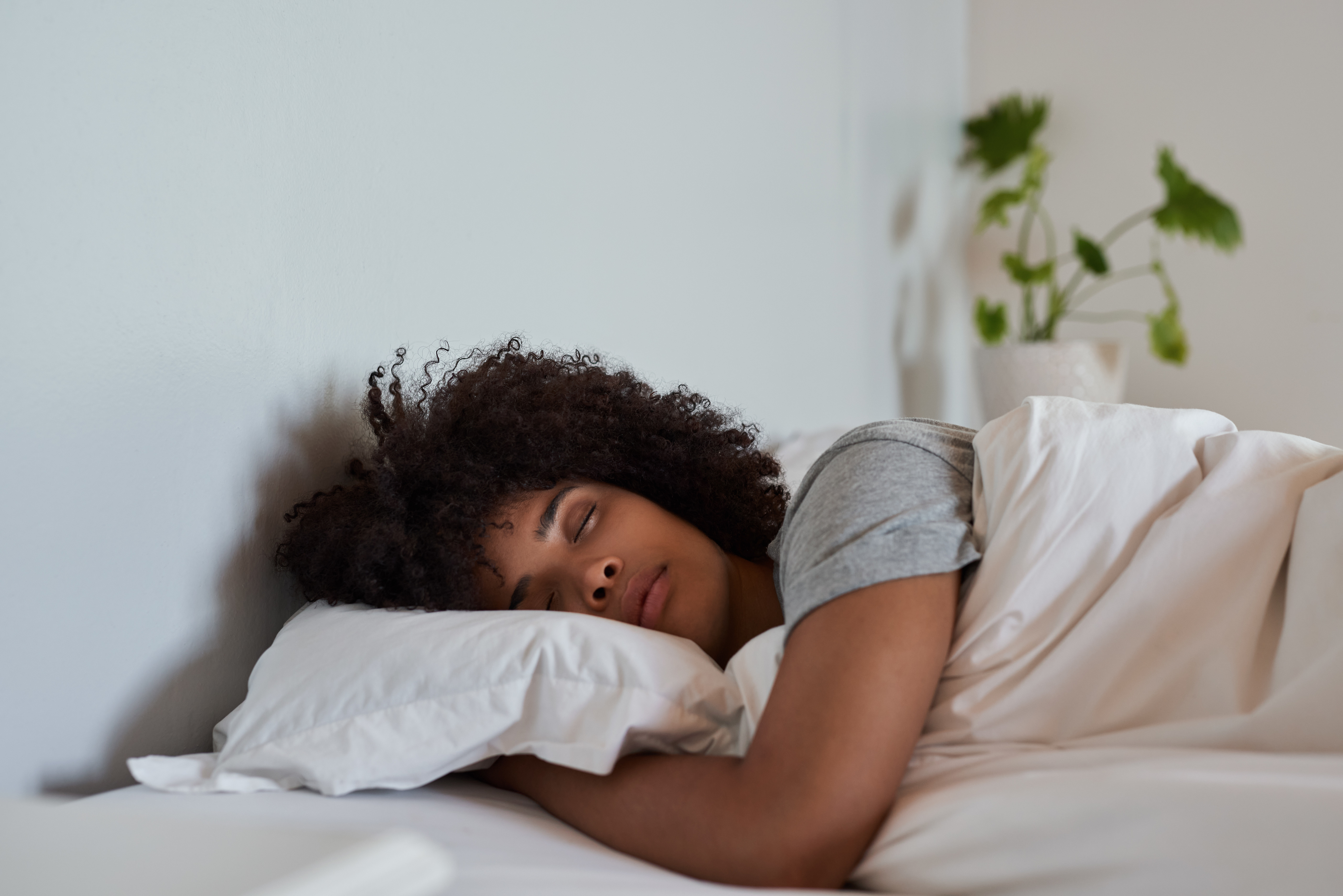 A person with curly hair is sleeping peacefully in bed, lying on their side with their head resting on a pillow. A plant is visible in the background