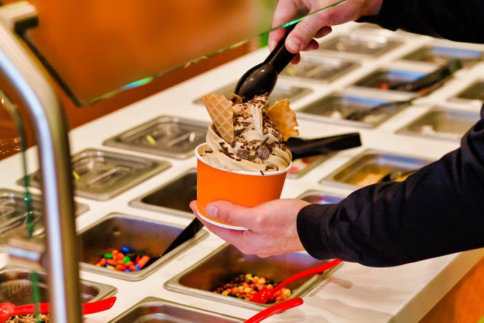 Person holding a cup filled with soft-serve ice cream and various toppings over a toppings bar station