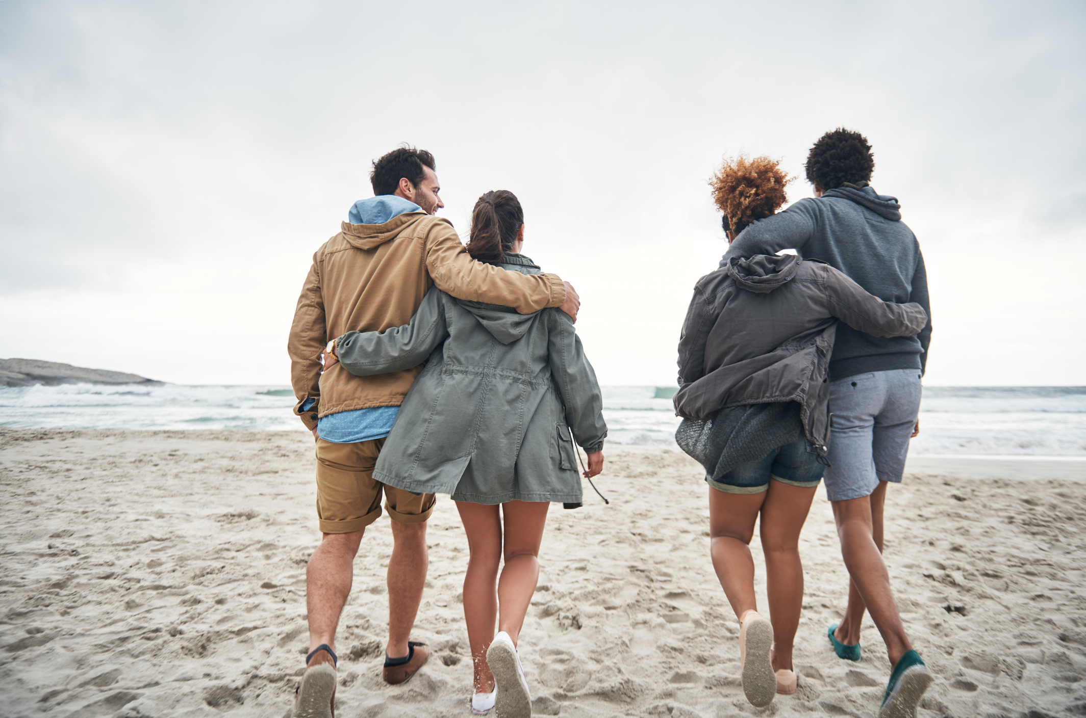 Four people walking on a beach with their arms around each other's shoulders, facing the ocean. They are wearing casual jackets and shorts. Their identities are unknown