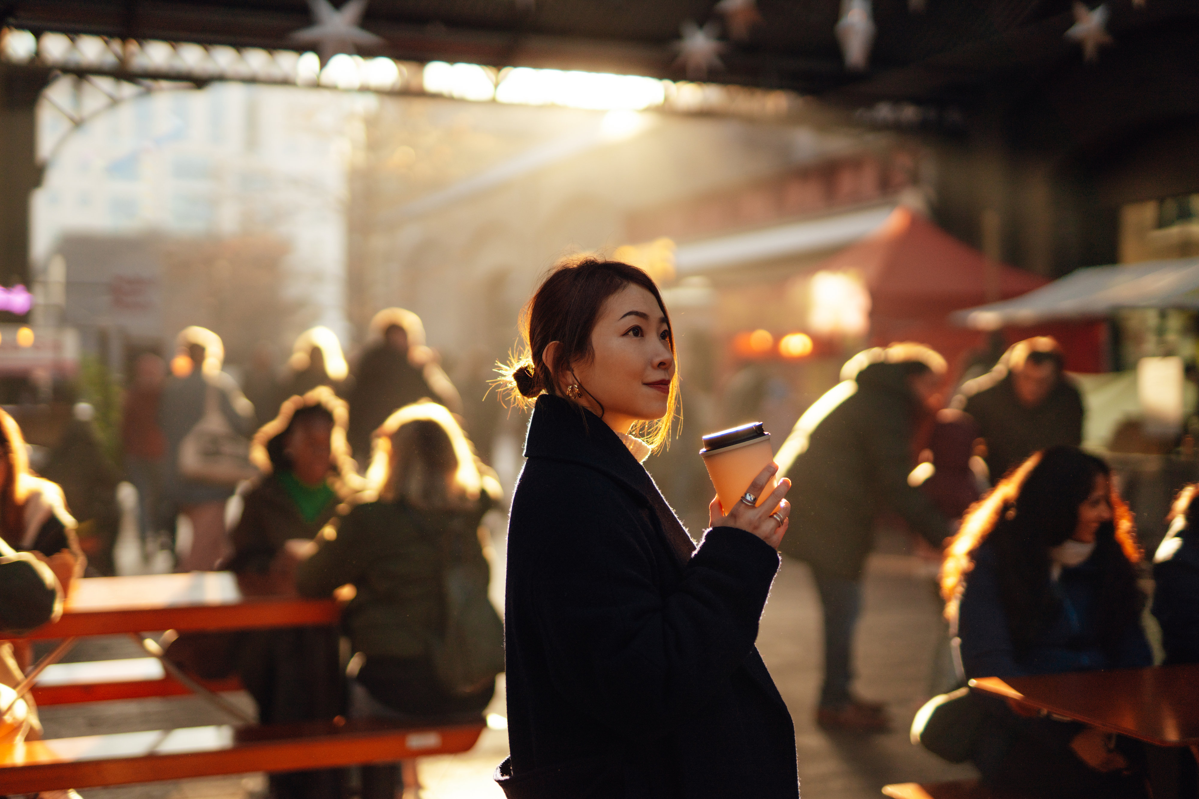 A woman in a marketplace holds a coffee cup, bathed in soft sunlight, with blurred people and booths in the background
