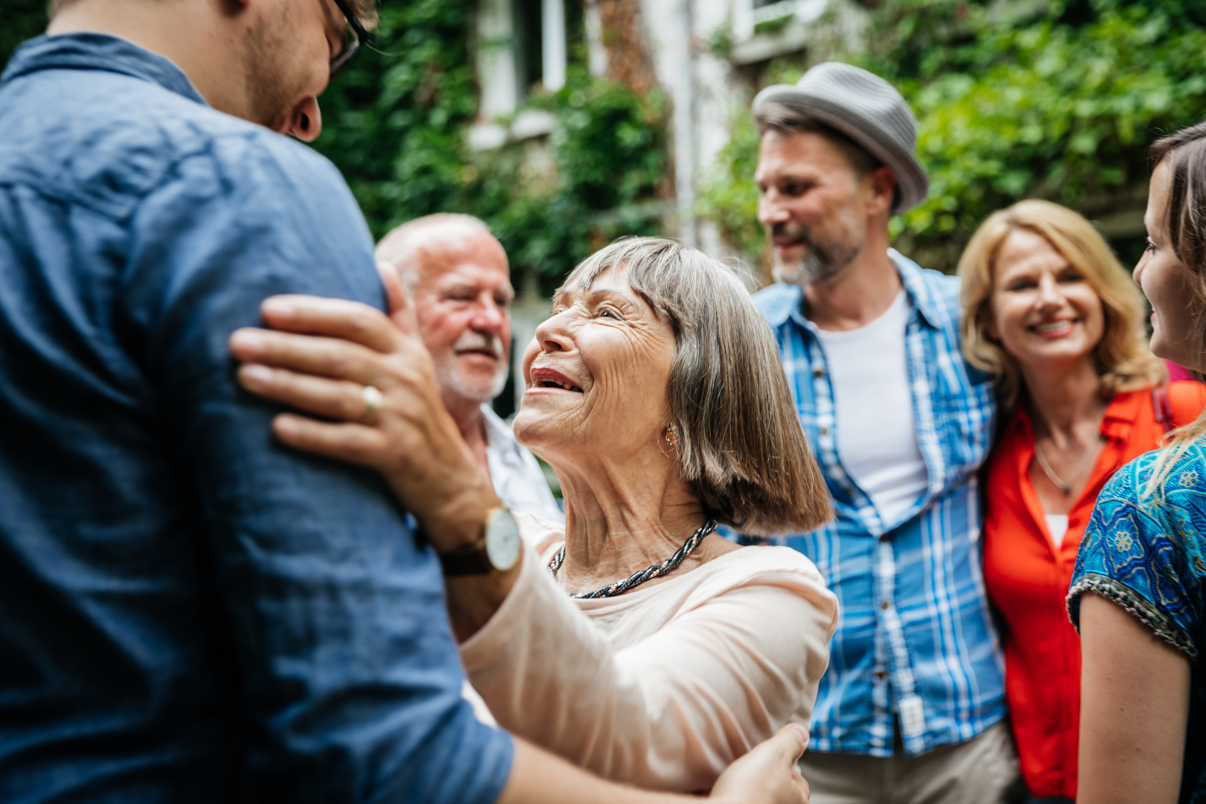 A group of people, including an elderly woman and others conversing and smiling, are gathered outside. The elderly woman is wearing a long-sleeved shirt and glasses