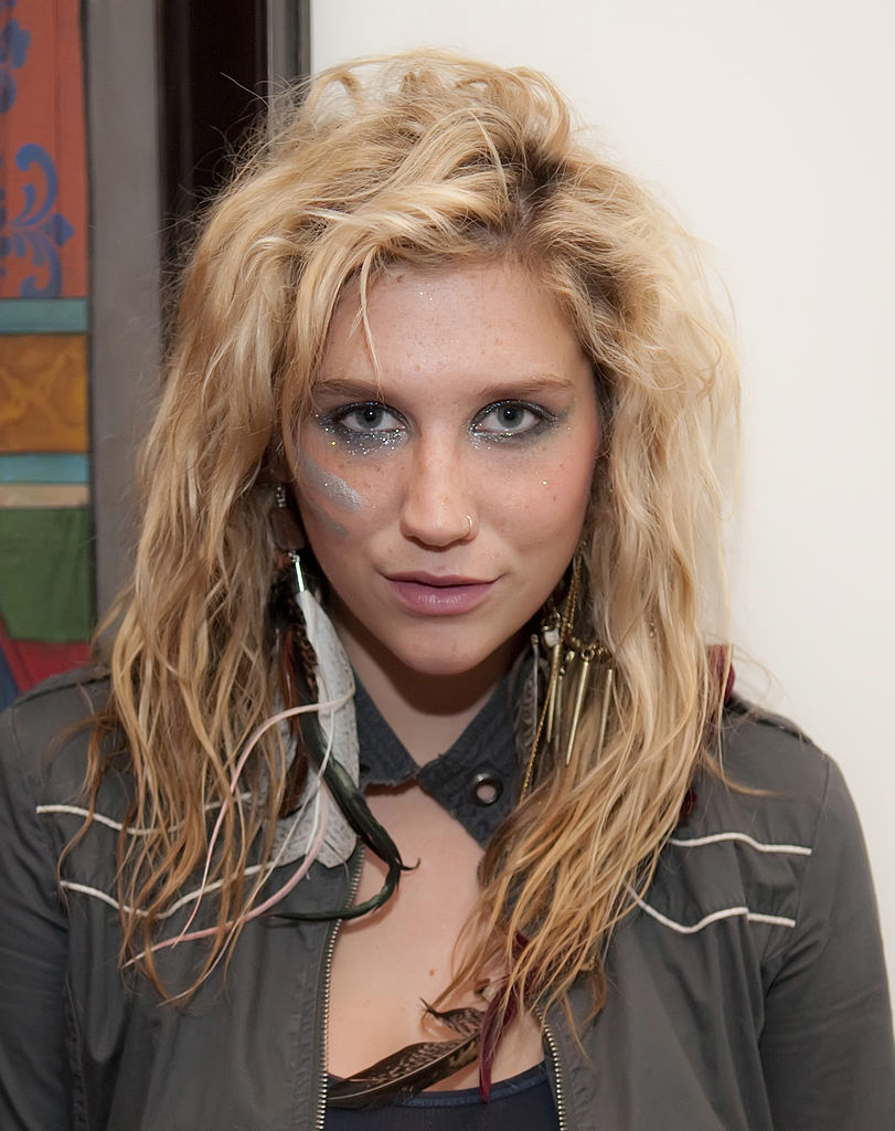 Kesha with wavy hair and glittery makeup, wearing a casual jacket and long feather earrings at an event