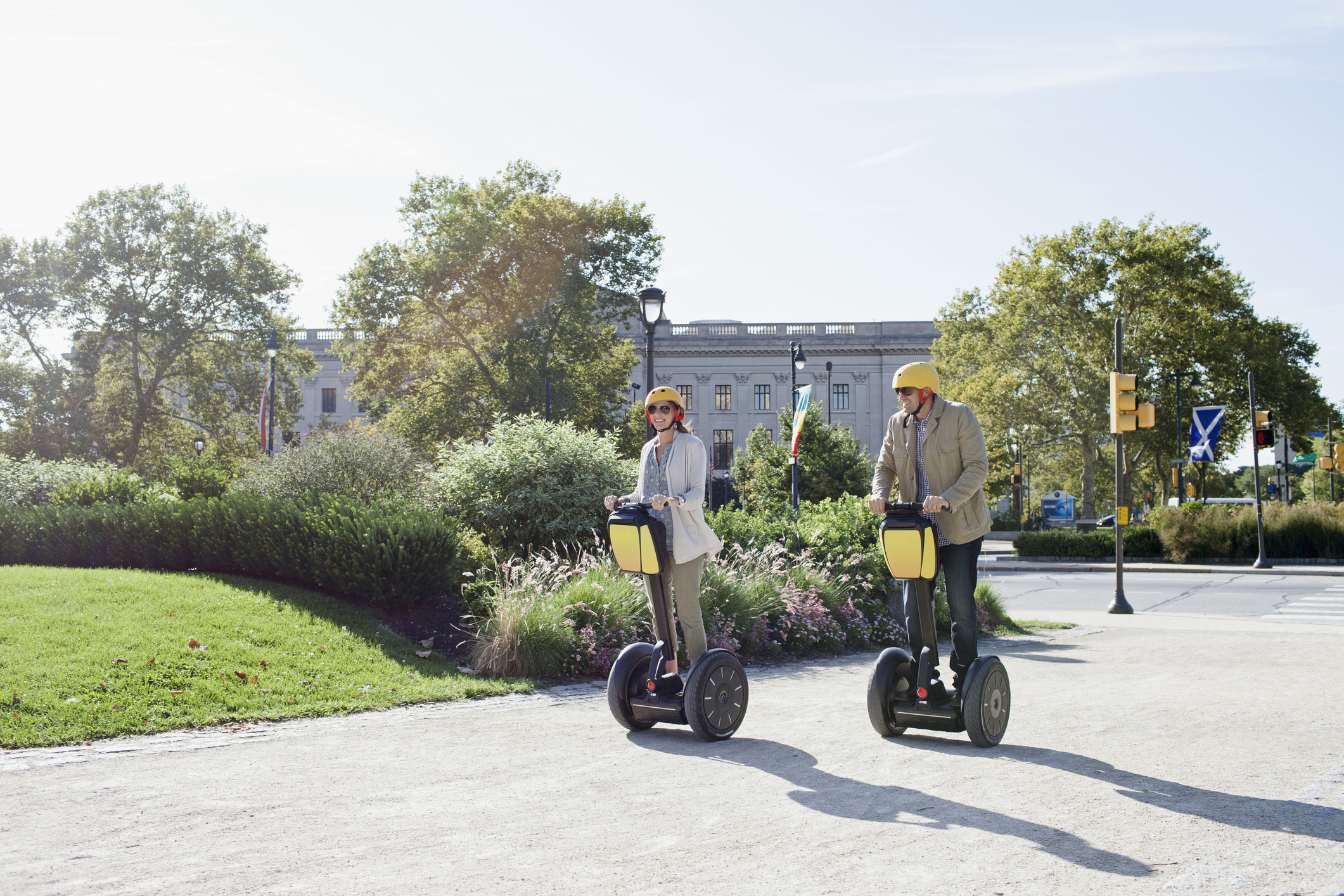 Two people wearing helmets ride Segways in a sunny park with greenery and buildings in the background