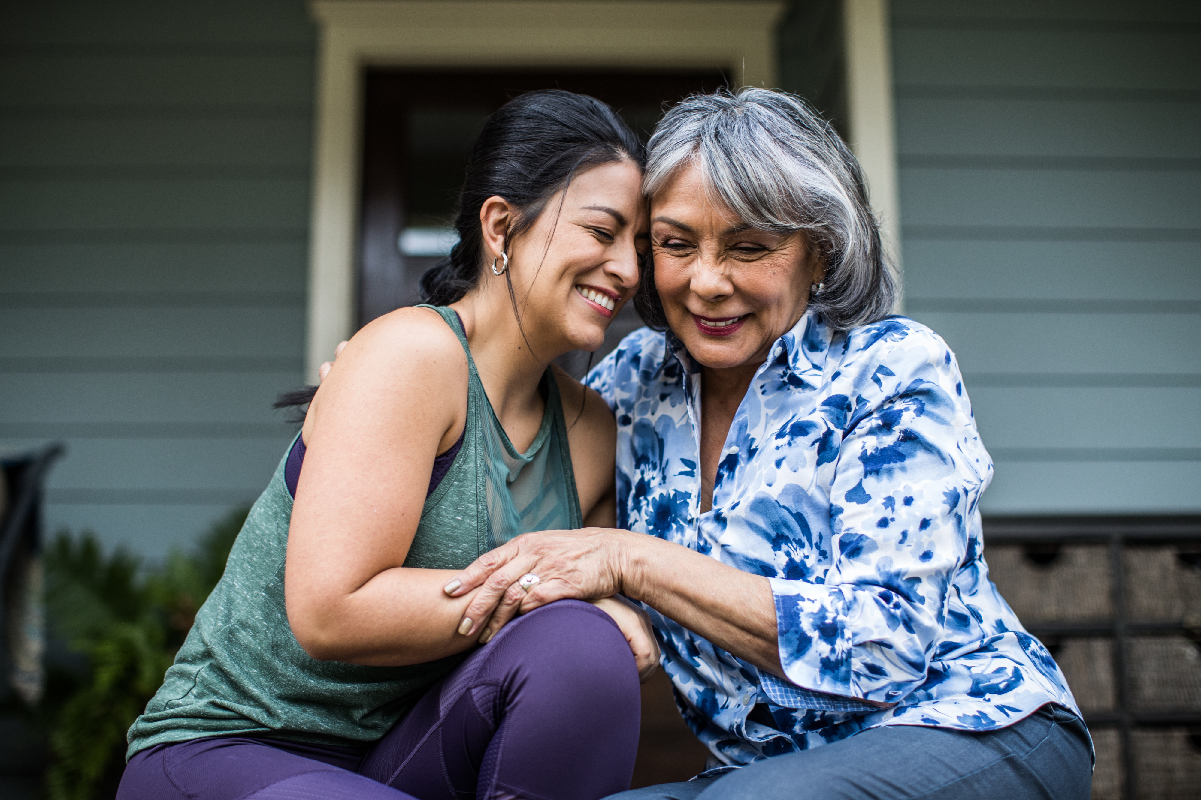 Two women share a warm hug while sitting on a porch step. The older woman wears a patterned blouse, and the younger woman wears a casual top and leggings